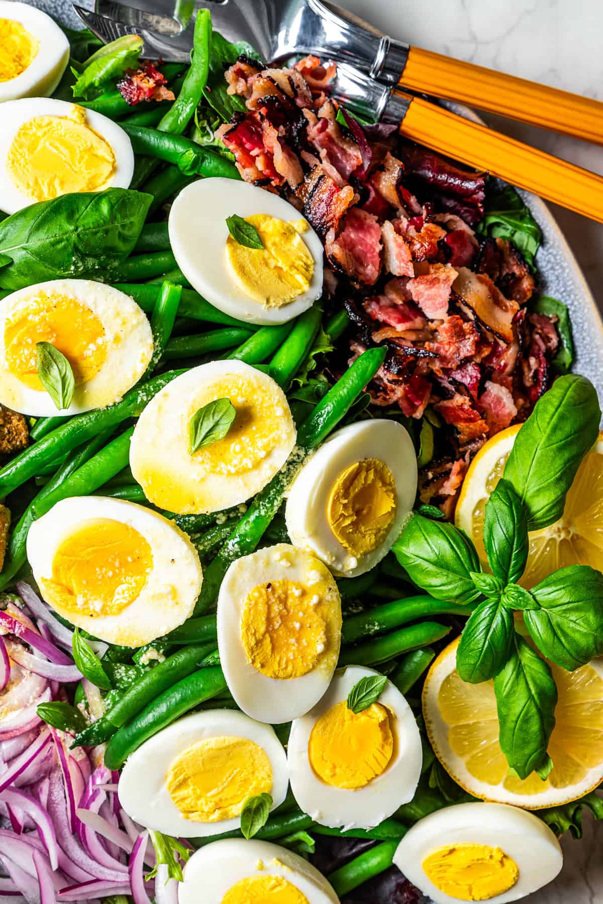 Green bean salad on a large platter topped with boiled eggs, crumbled bacon, and fresh basil.