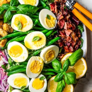 A large platter with green beans, halved boiled eggs, crumbled bacon, croutons, red onions, sliced lemons, and a garnish of fresh basil leaves.