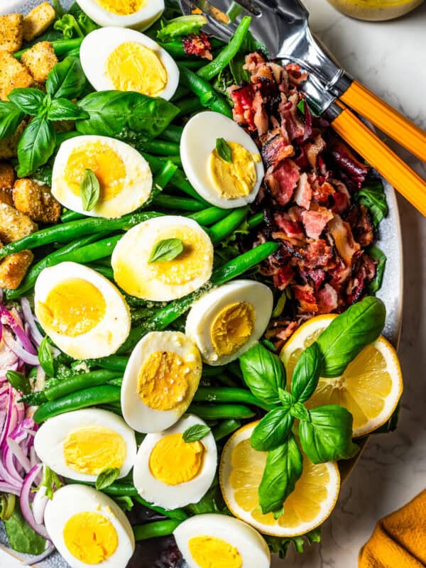 A large platter with green beans, halved boiled eggs, crumbled bacon, croutons, red onions, sliced lemons, and a garnish of fresh basil leaves.