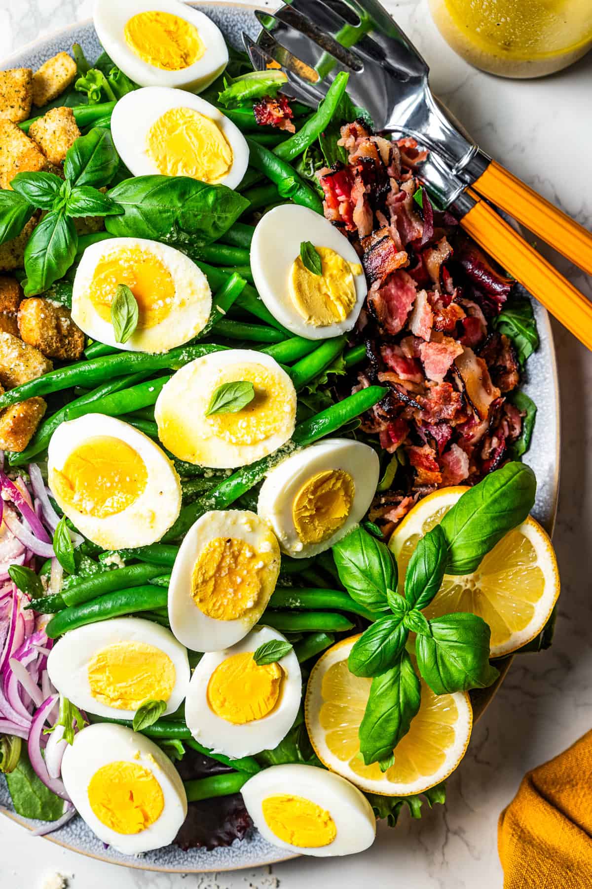 A large platter with green beans, halved boiled eggs, crumbled bacon, croutons, red onions, sliced lemons, and a garnish of fresh basil leaves.