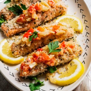 Panko-crusted salmon filets served on a platter next to lemon wedges and topped with Tuscan tomato sauce.