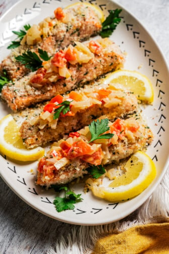 Panko-crusted salmon filets served on a platter next to lemon wedges and topped with Tuscan tomato sauce.
