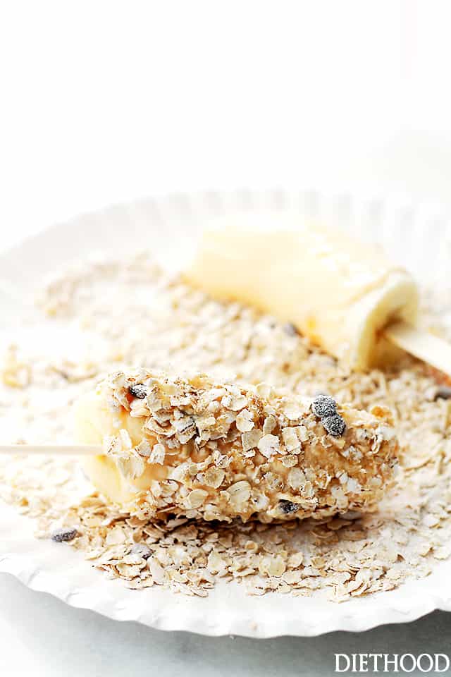 Banana popsicles being coated with granola on a paper plate.
