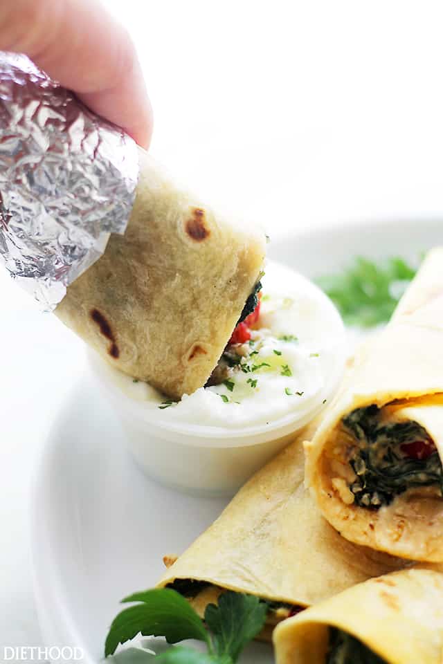 Esta fácil envoltura de queso feta de espinacas tiene un relleno cremoso de pimientos salteados, cebollas, espinacas y queso enrollados y horneados dentro de una tortilla tibia. Una mano que sumerge una envoltura de queso espinacas en un pequeño tazón de salsa para mojar junto a más envolturas en un plato.