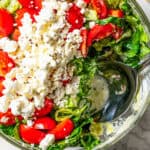 Mediterranean seven layer dip in a glass bowl with a serving spoon resting inside the bowl.