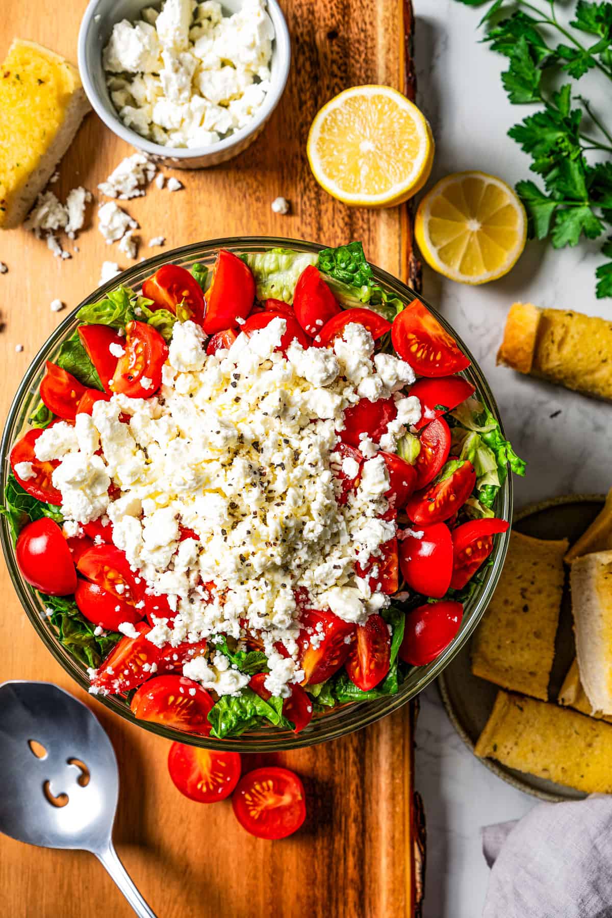 Overhead view of Mediterranean seven layer dip in a glass bowl next to two lemon halves and a small bowl of crumbled feta cheese.