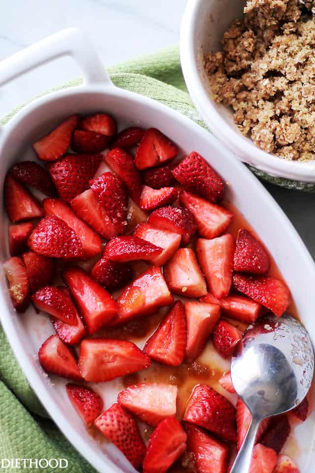 Chopped strawberries spread into the bottom of an oval baking dish with a spoon, next to a bowl of oat crumble topping.