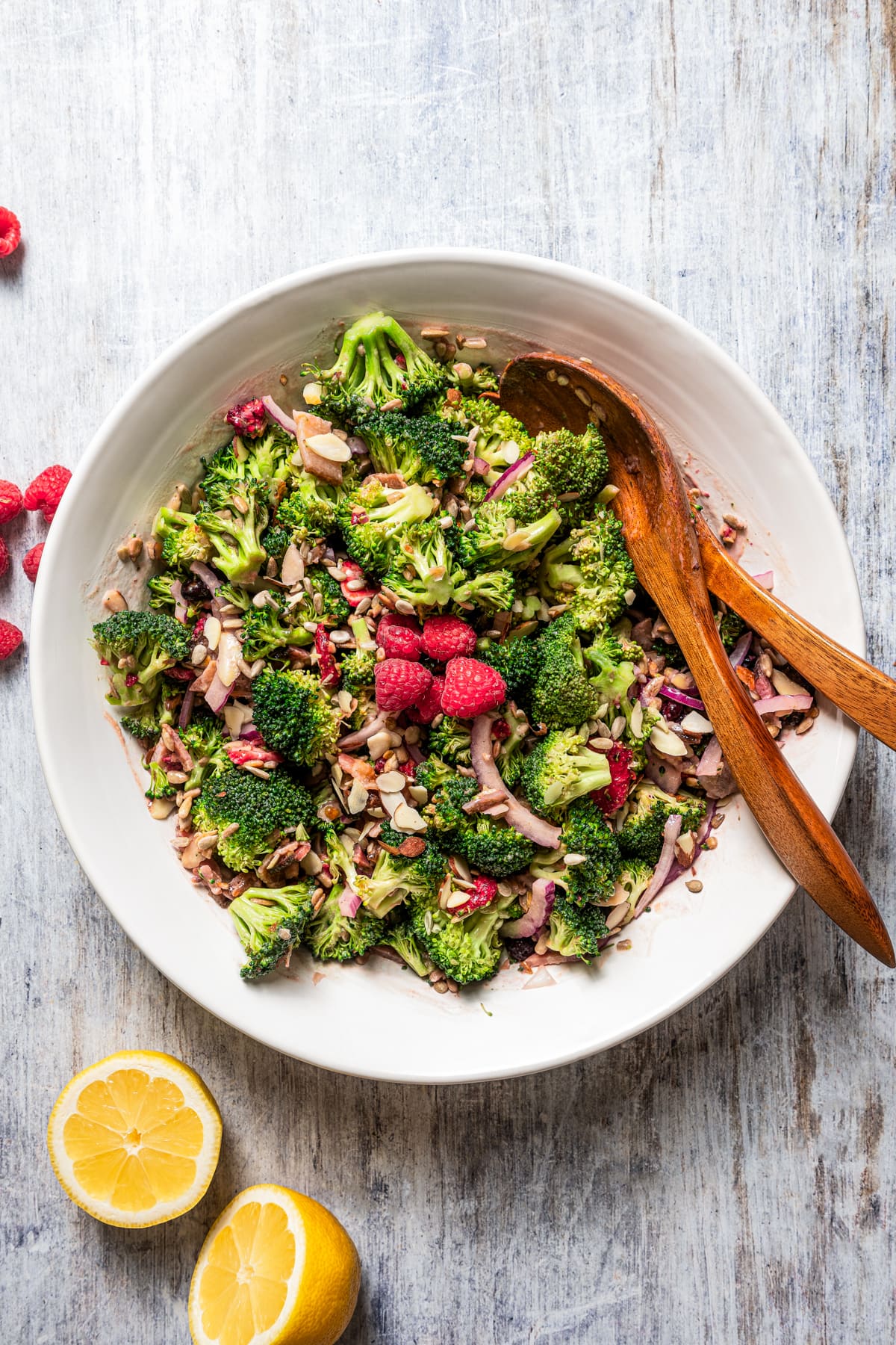 Overhead image of broccoli salad in a salad bowl with serving spoons and fork and a few fresh raspberries for garnish.