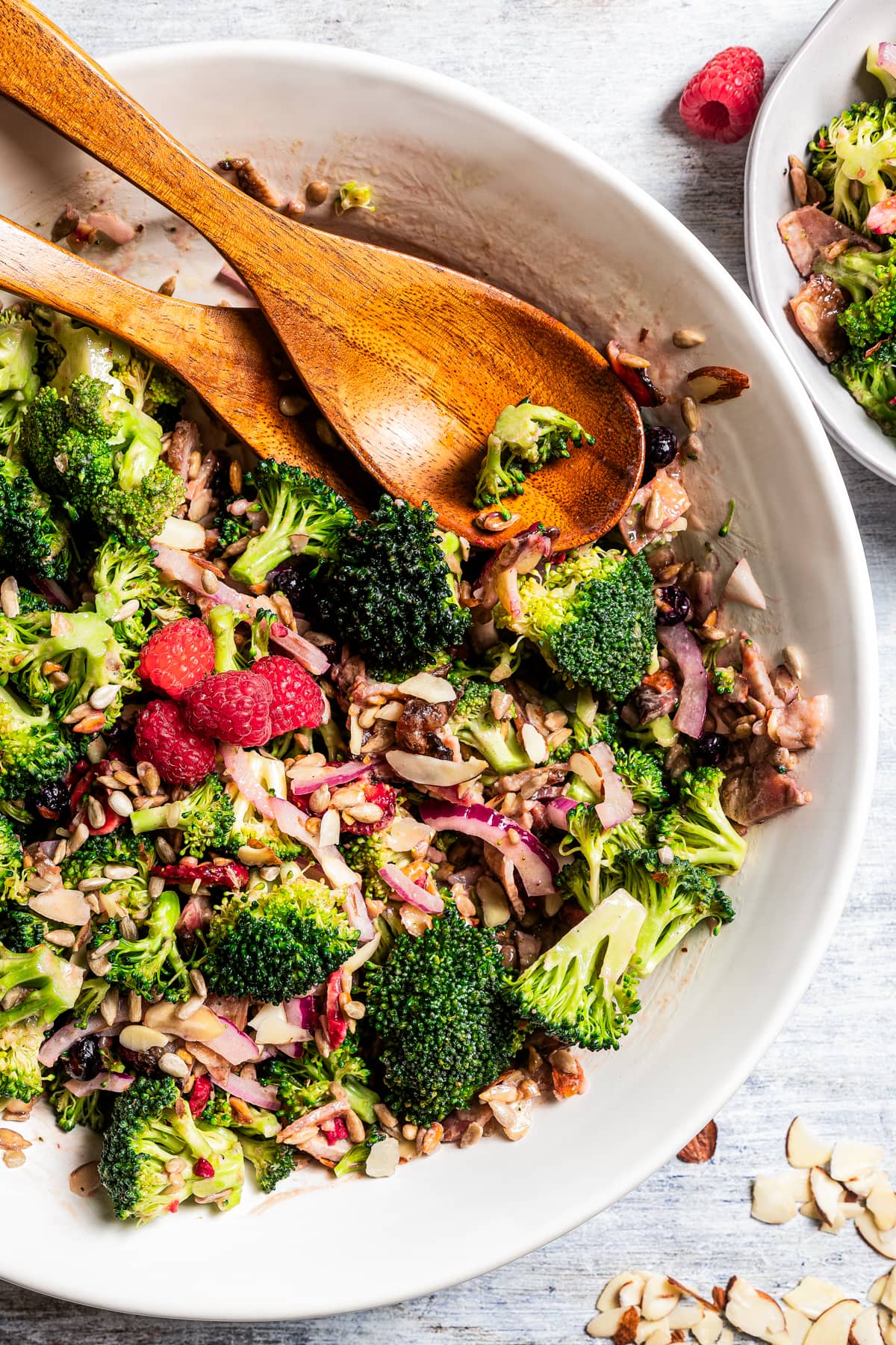 Broccoli salad in a serving bowl with salad tongs and a couple of fresh raspberries.