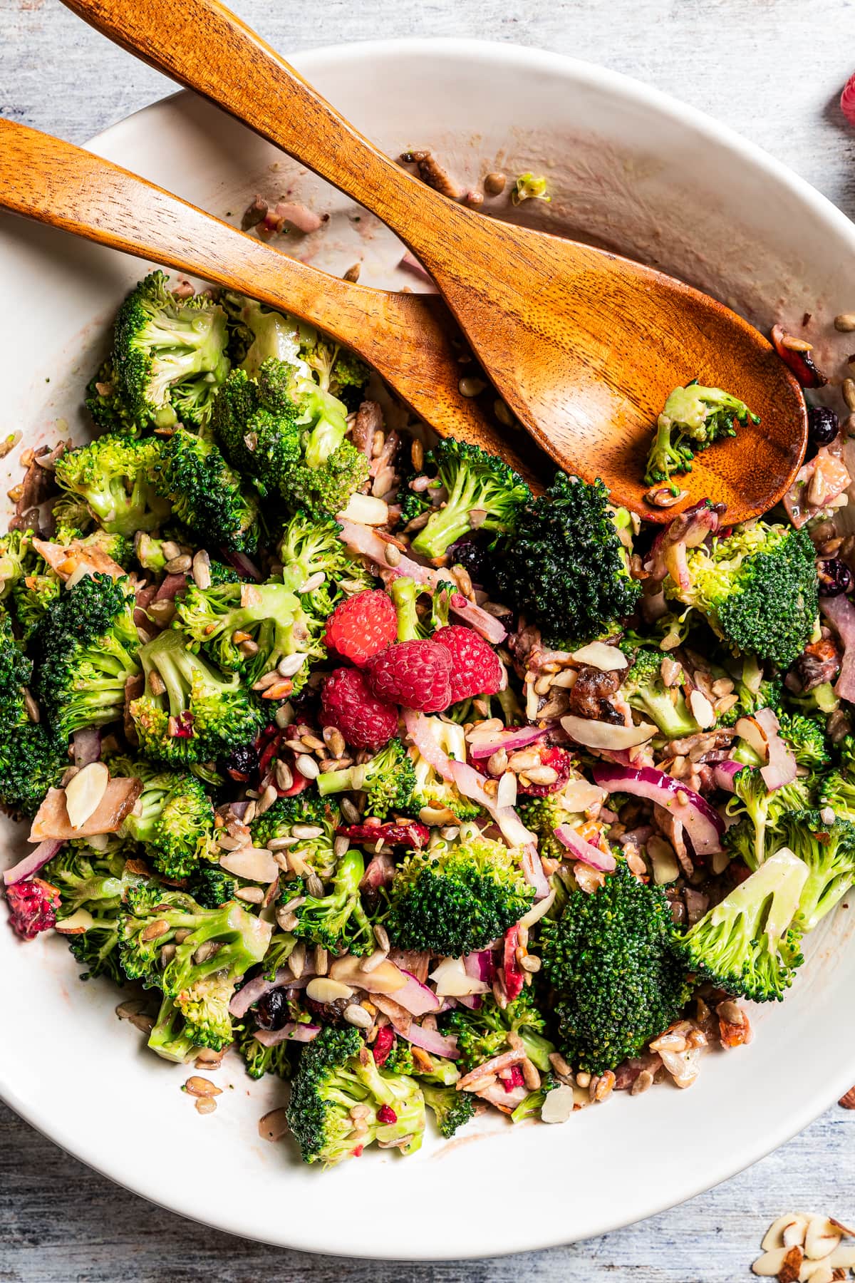 Broccoli salad in a serving bowl with salad tongs and a couple of fresh raspberries.