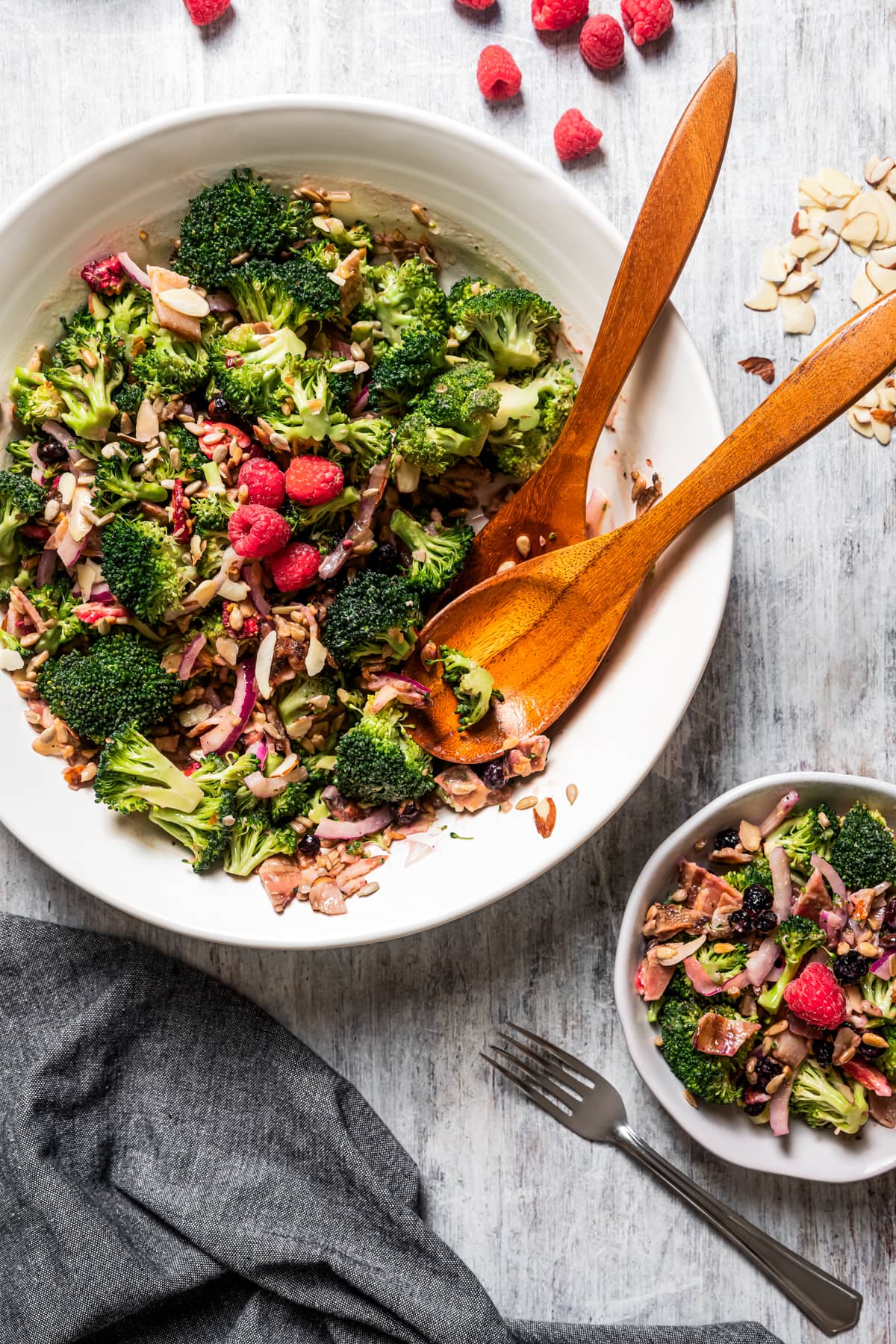 Overhead image of broccoli salad in a serving bowl with tongs and a couple of fresh raspberries. A halved lemon sits nearby.