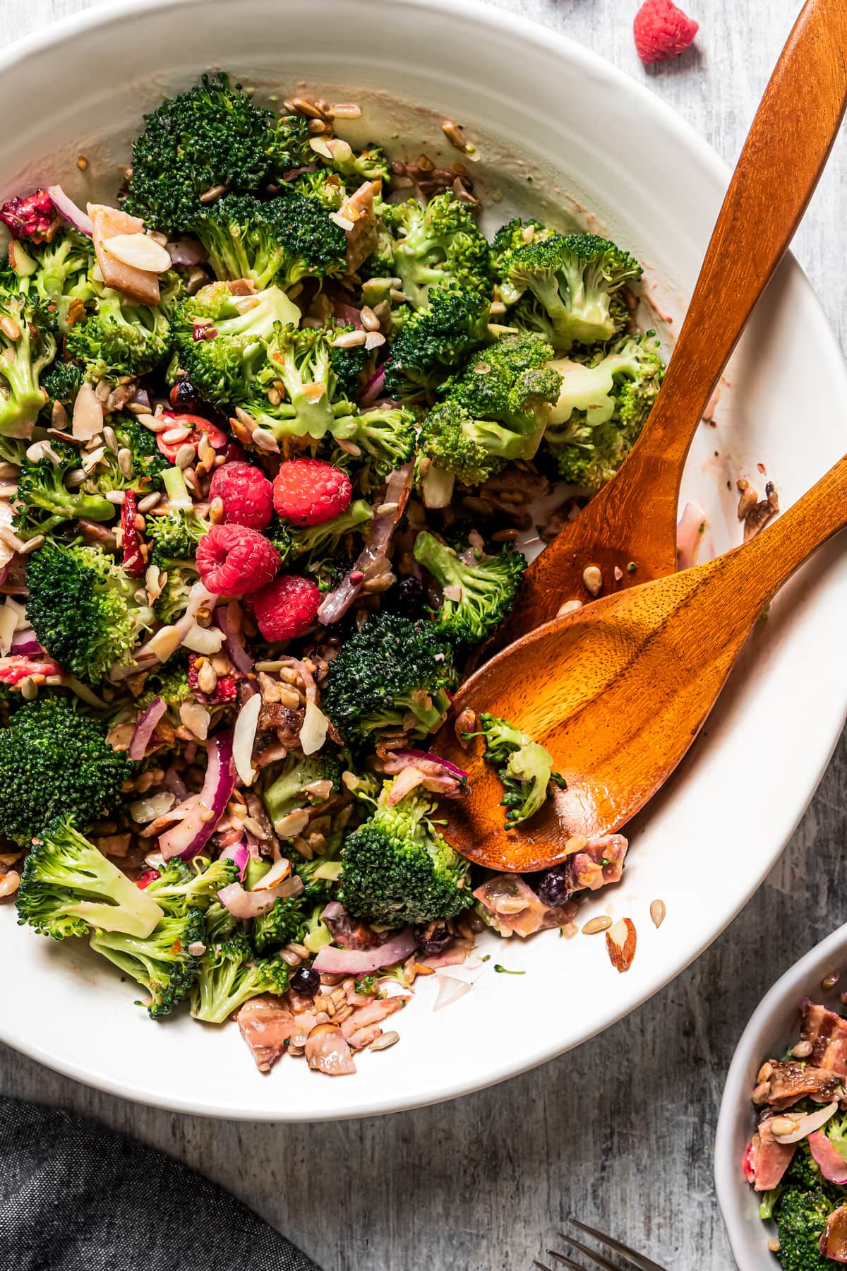 Broccoli salad in a serving bowl with salad tongs and a couple of fresh raspberries.