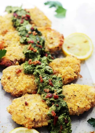 Beautifully crusted pork chops lined up on a plate with pesto on top that has visible bacon crumbled in it.