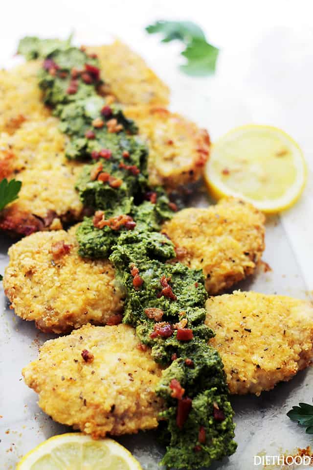 Beautifully crusted pork chops lined up on a plate with pesto on top that has visible bacon crumbles in it.
