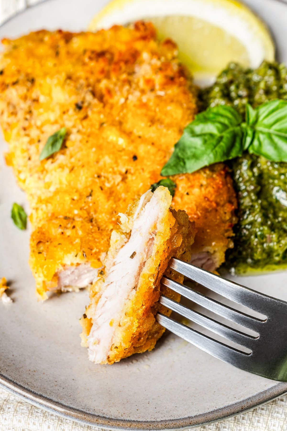 A fork taking a bite out of breaded pork chops served on a plate with bacon-kale pesto and a slice of lemon.