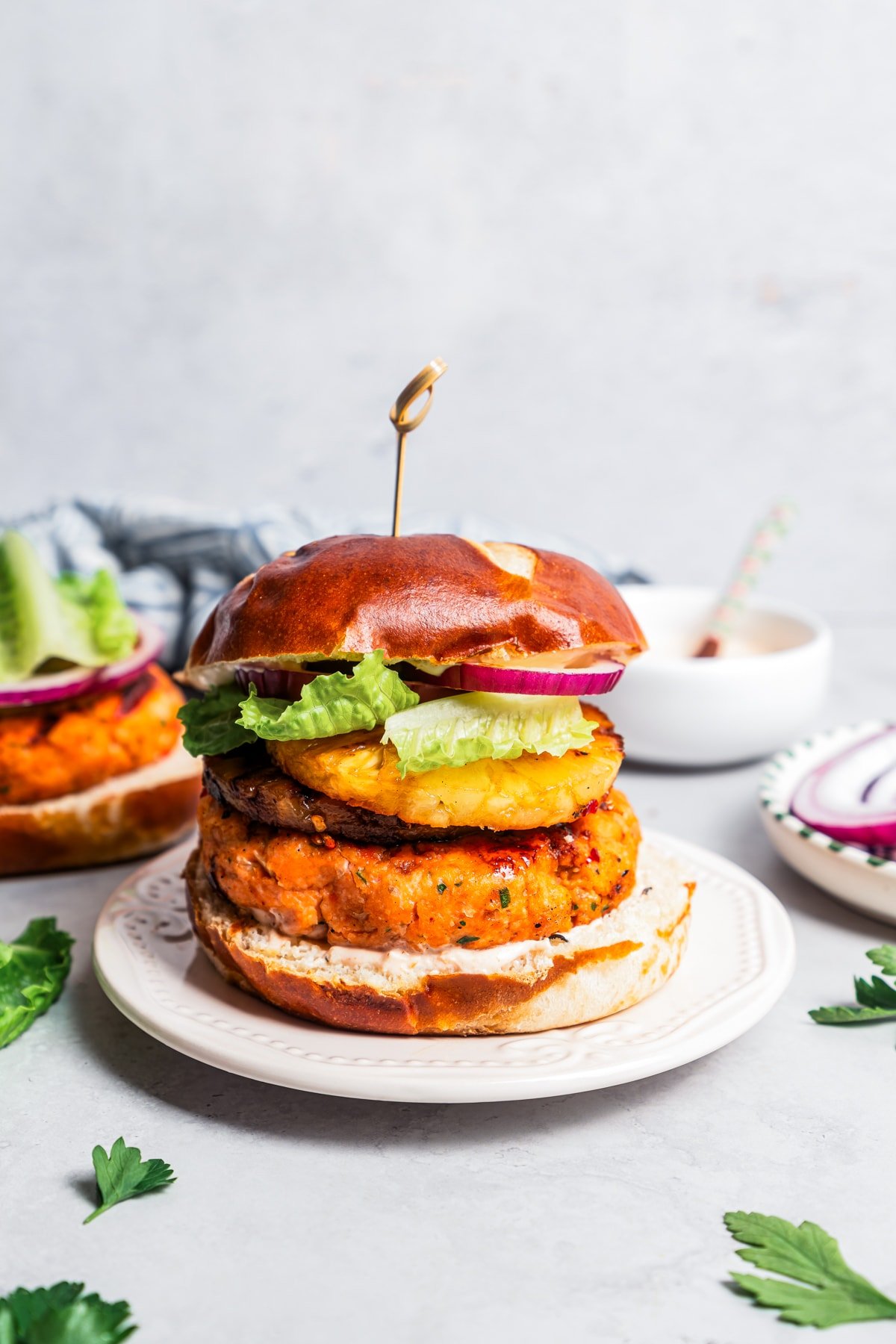 A grilled salmon burger on a plate with another behind it being topped with red onion and lettuce.