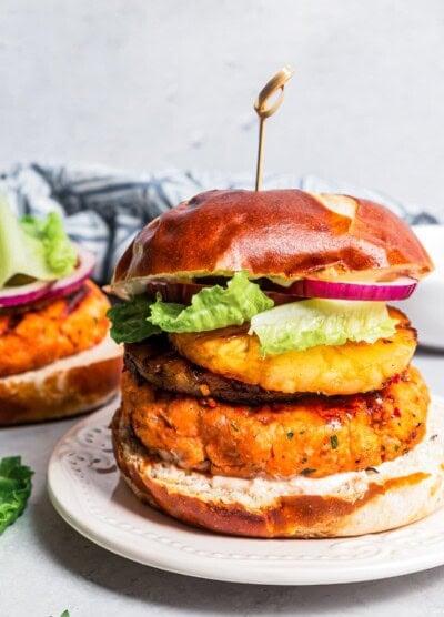 A grilled salmon burger on a plate with another behind it being topped with red onion and lettuce.