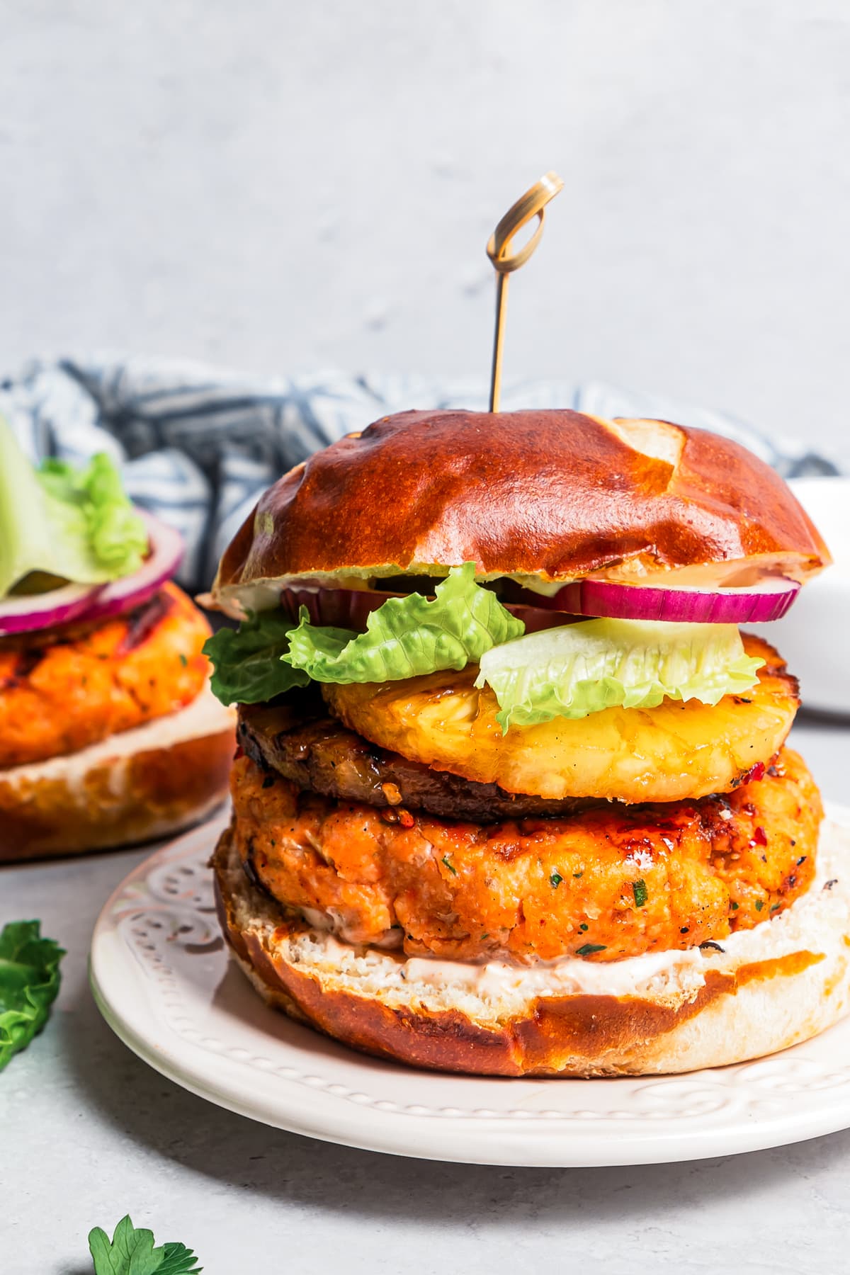 A grilled salmon burger on a plate with another burger behind it being topped with red onion and lettuce.