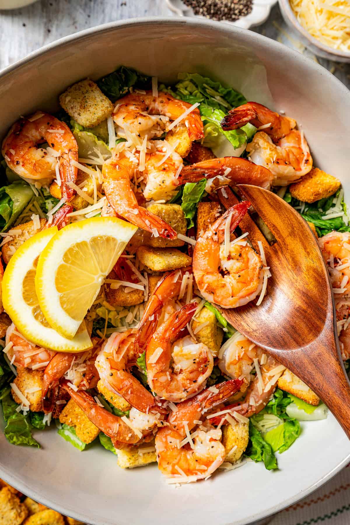 Overhead view of shrimp Caesar salad in a large bowl garnished with lemon wedges next to a wooden salad spoon.