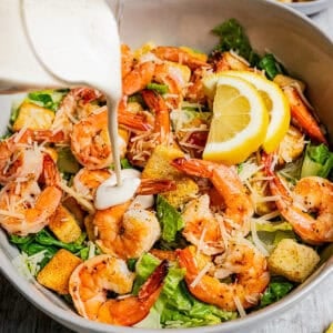 Pouring Caesar salad dressing over shrimp and lettuce in a bowl.