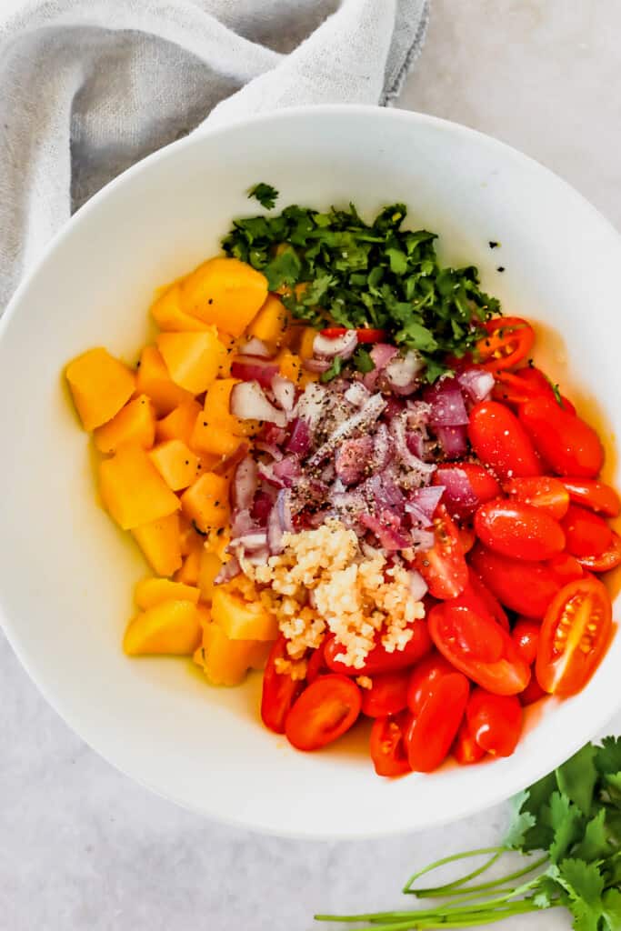 white bowl with chopped mangoes, halved cherry tomatoes, red onions, and green herbs.
