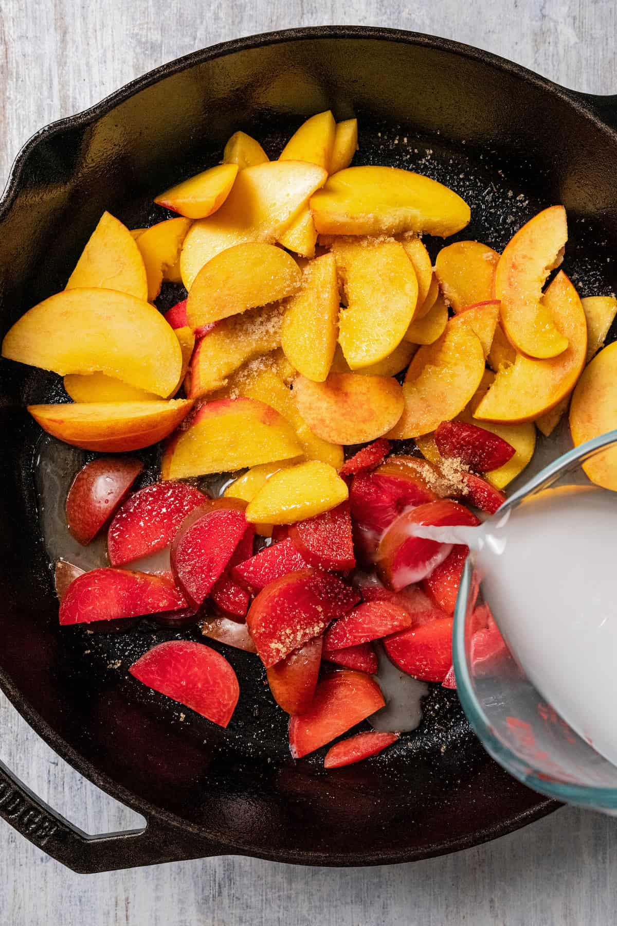 A cornstarch slurry being poured over peach and plum slices in a skillet.