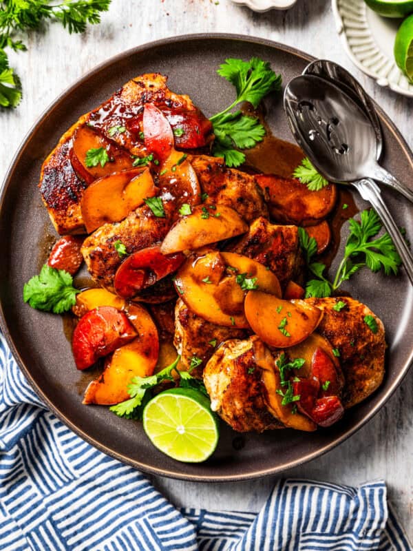 An overhead view of Jamaican jerk chicken topped with stone fruit, including peaches and plums, is served on a plate garnished with half a lime next to two serving spoons.