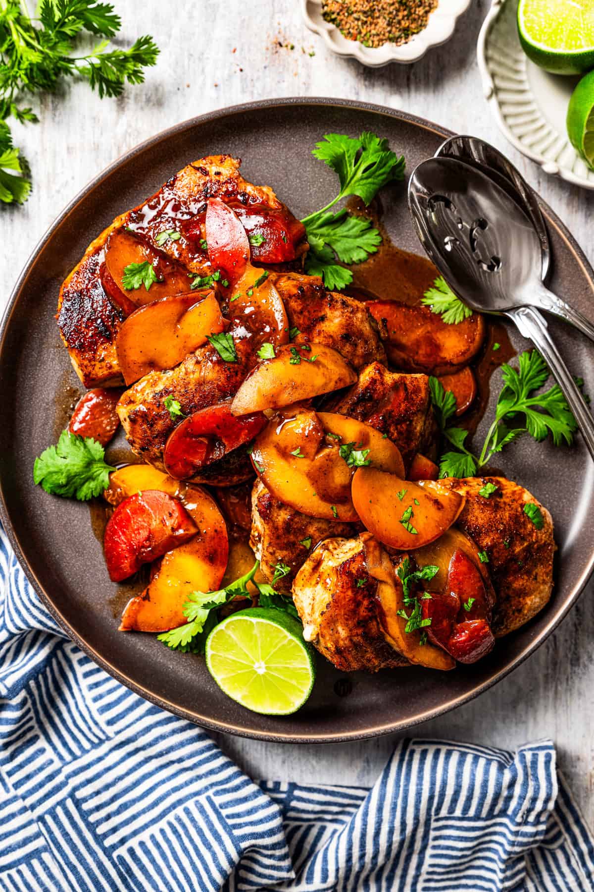 An overhead view of Jamaican jerk chicken topped with stone fruit, including peaches and plums, is served on a plate garnished with half a lime next to two serving spoons.