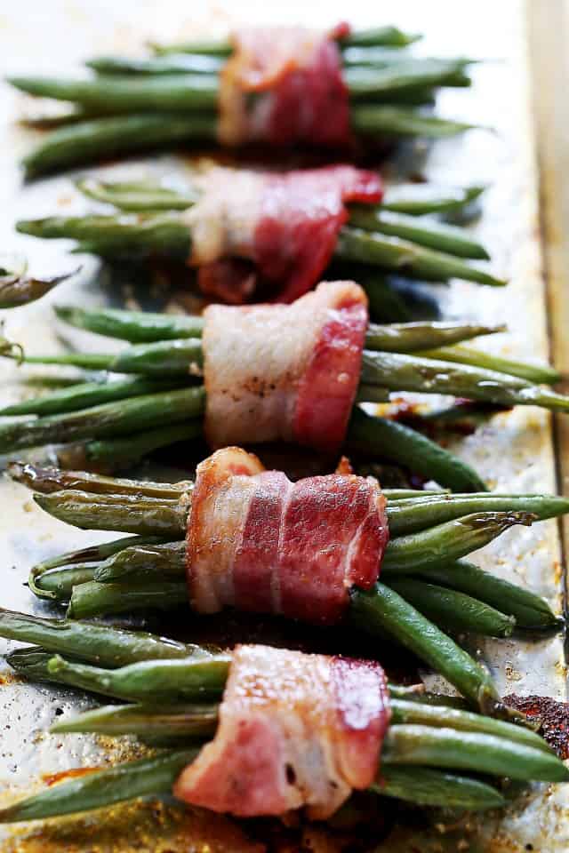 A row of five bundles of green beans wrapped in crispy bacon, on a baking sheet. 