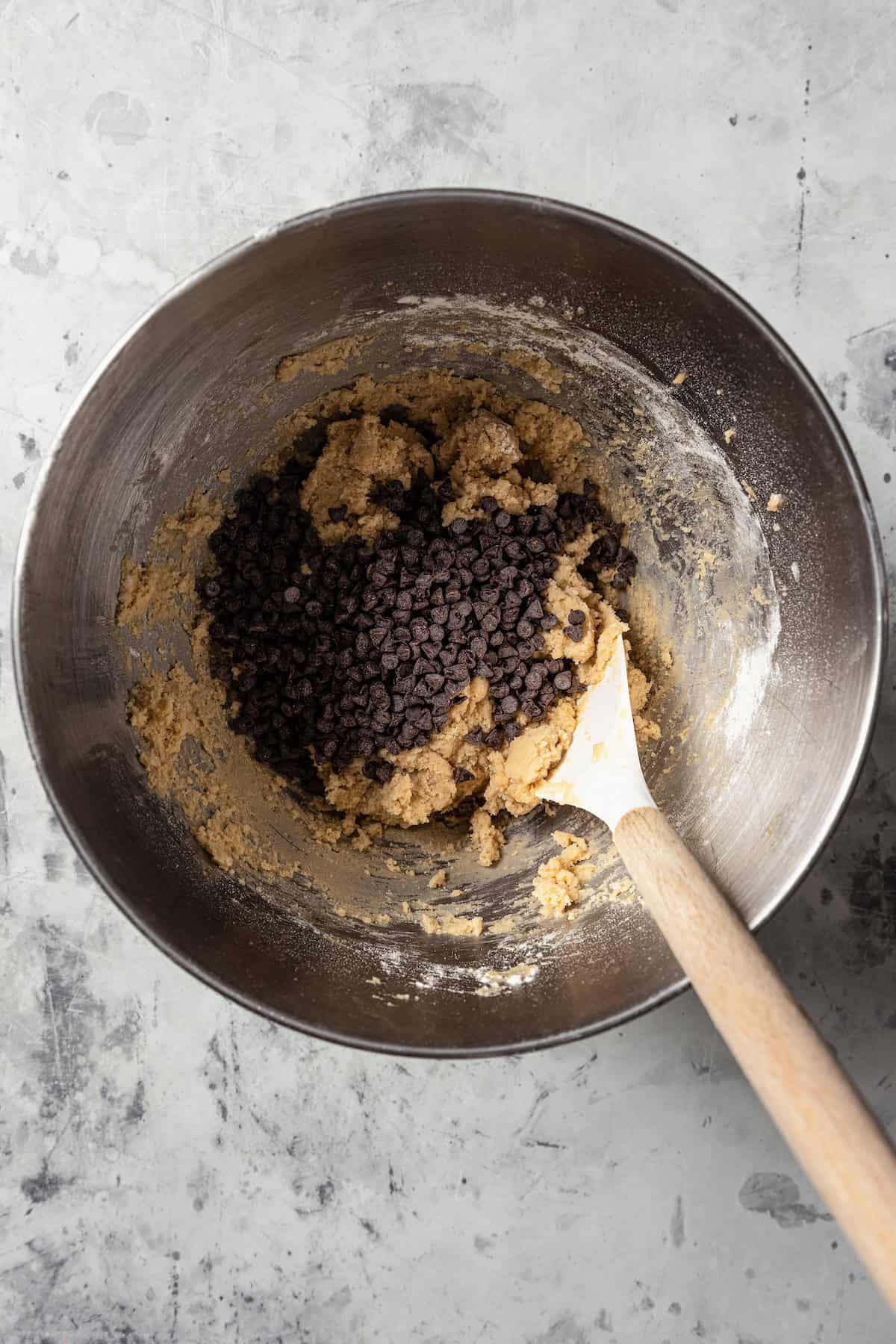 Mini chocolate chips added to cookie dough in a metal mixing bowl.