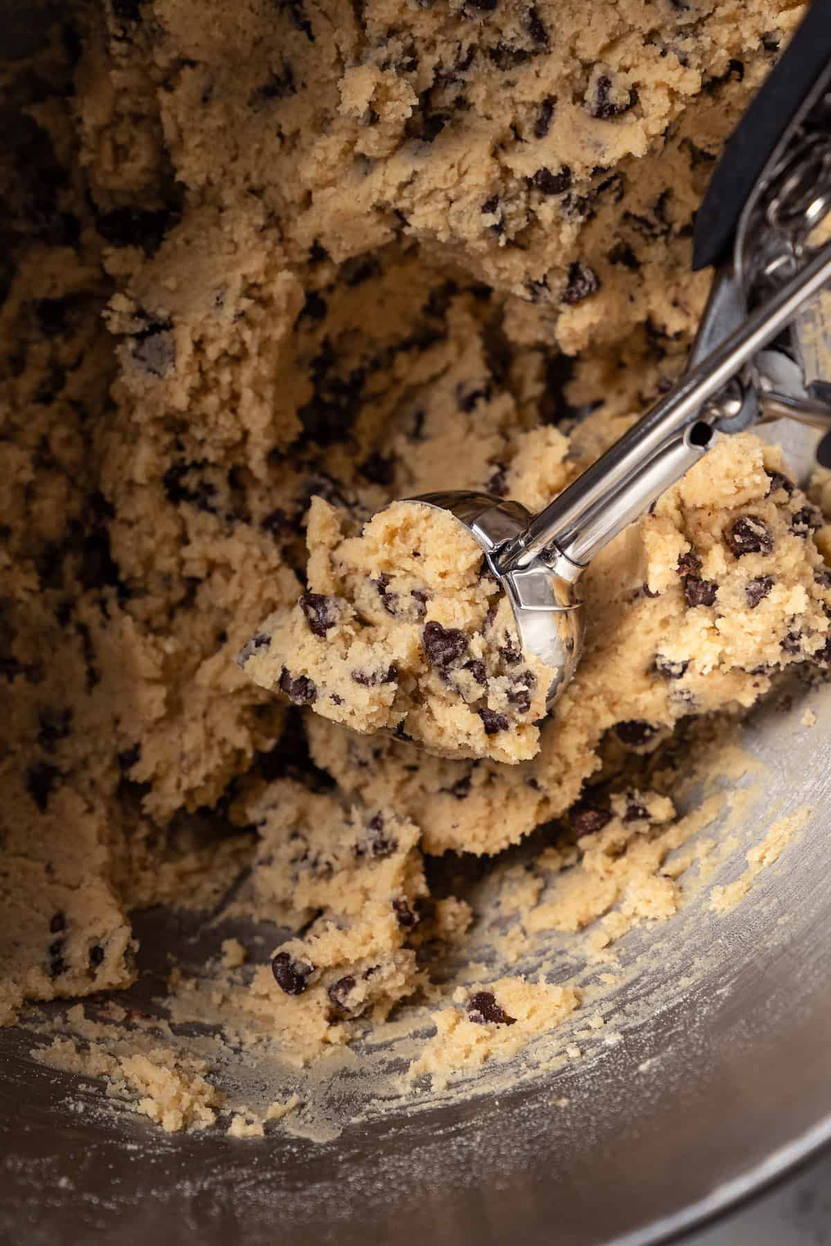 A cookie scoop scooping chocolate chip cookie from a metal mixing bowl.