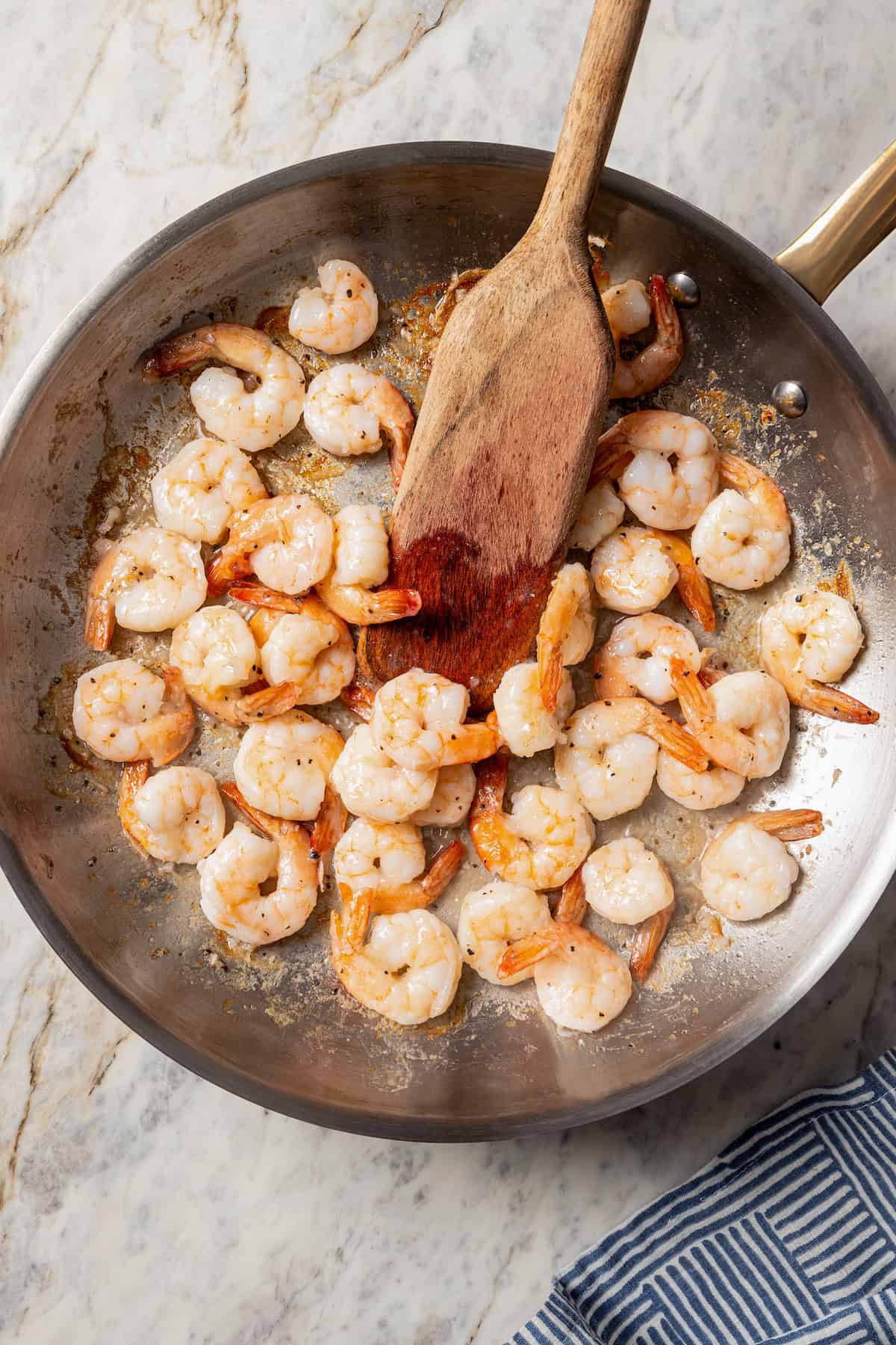 A wooden spoon stirring fried shrimp in a stainless steel skillet.