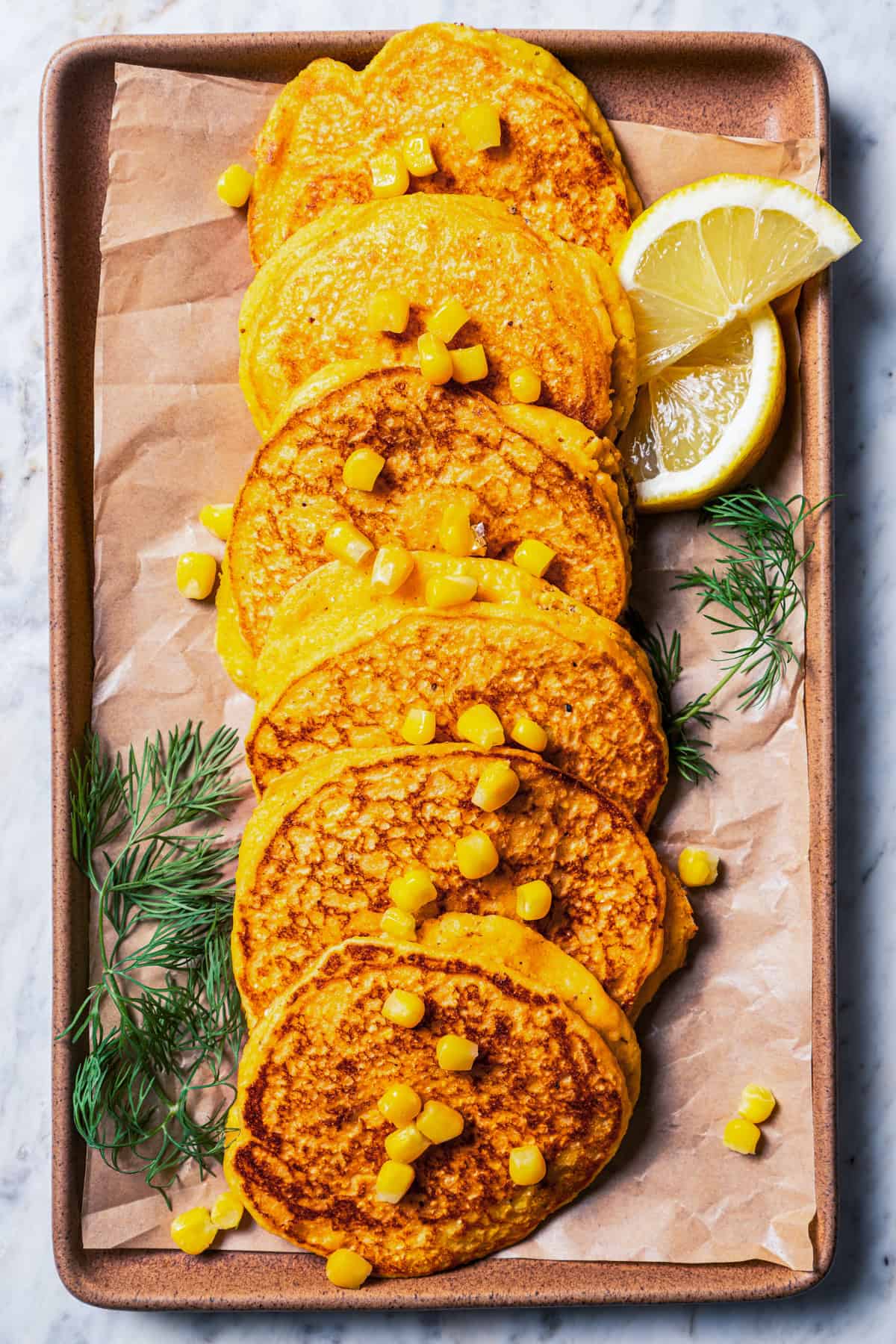 Cornbread pancakes arranged in a row on a parchment-lined tray, garnished with fresh dill, sweet corn kernels, and lemon wedges.