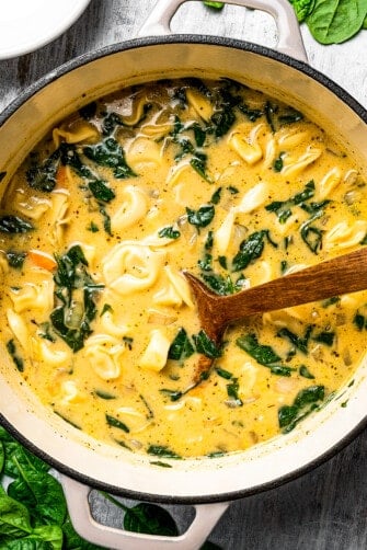 Creamy tortellini soup ingredients simmering in a pot with a wooden spoon.