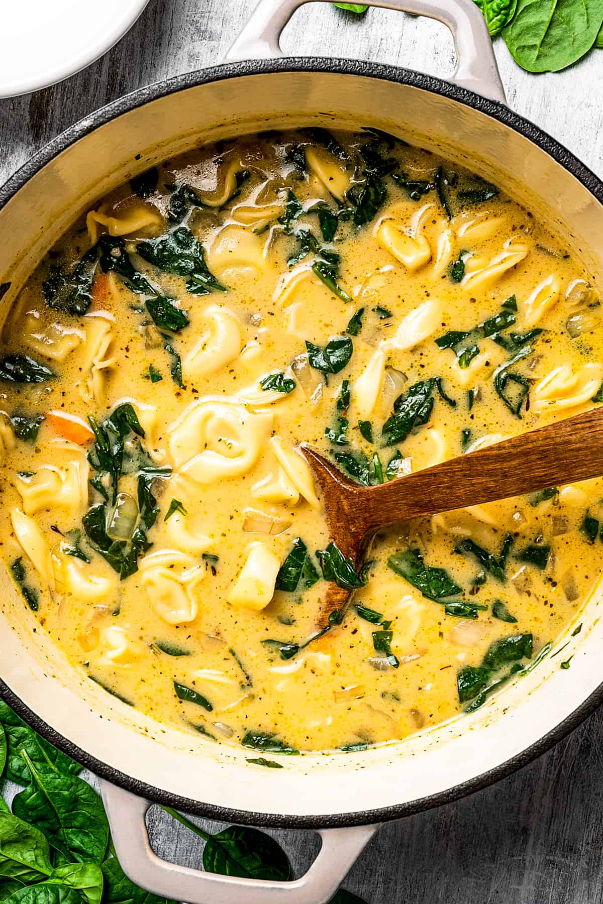 Creamy tortellini soup ingredients simmering in a pot with a wooden spoon.