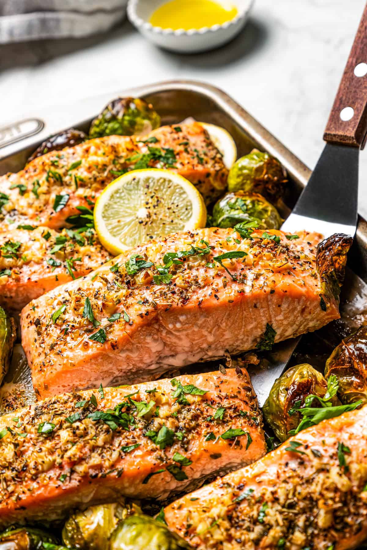 A spatula lifting a baked salmon fillet from a sheet pan, surrounded by more fillets and roasted Brussels sprouts.