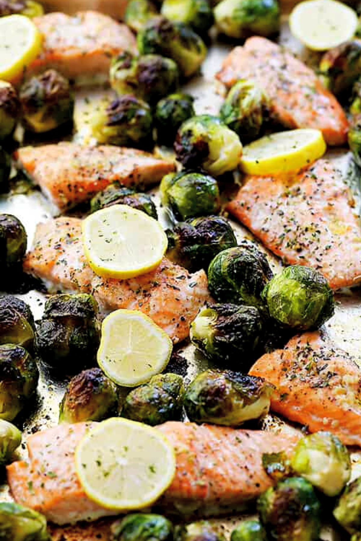 close up photo of a baking sheet with oven roasted salmon fillets topped with lemon slices, and brussel sprouts arranged around the fillets.