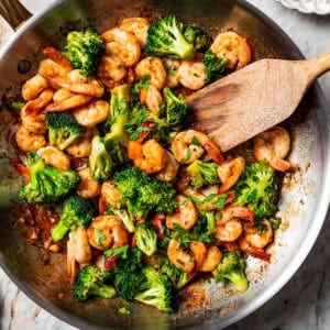 Wooden spoon stirring through shrimp stir fry in a skillet with broccoli.