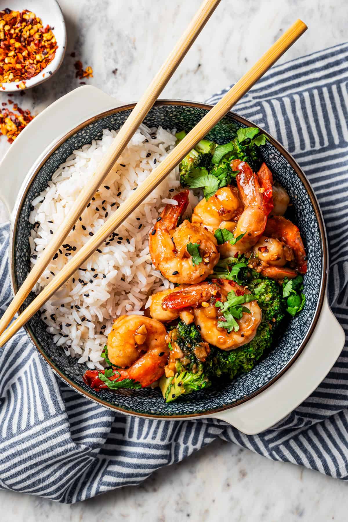 A pair of chopsticks resting on the side of a blue stoneware bowl filled with shrimp stir fry served over rice.