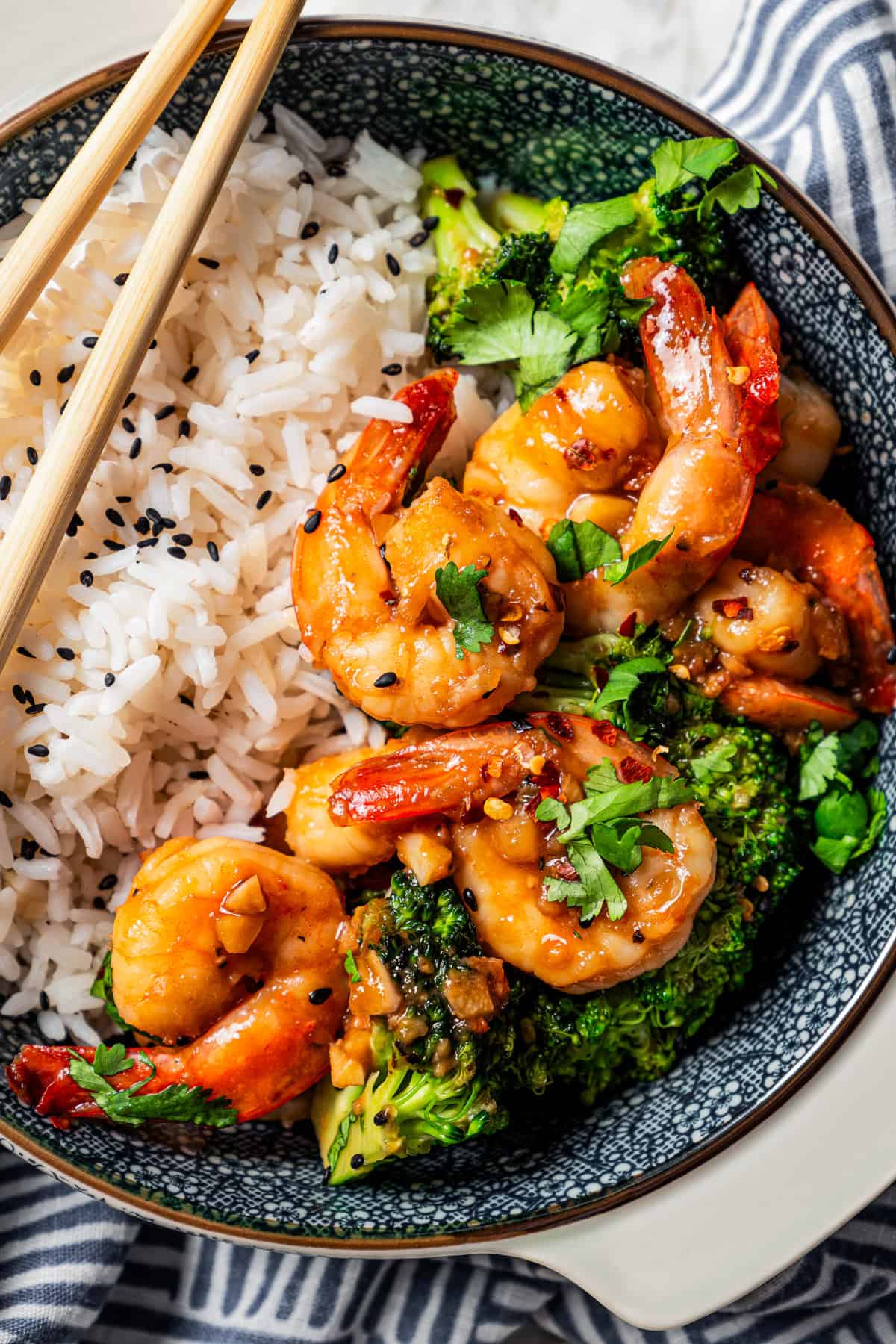 Close up of a pair of chopsticks resting on the side of a blue stoneware bowl filled with shrimp stir fry served over rice.