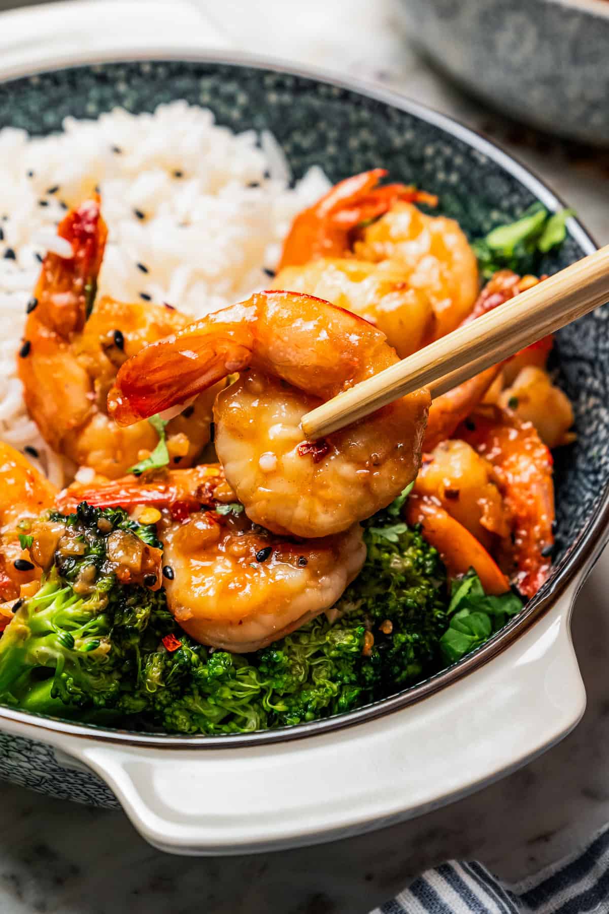 Chopsticks lifting a fried shrimp from a bowl of shrimp stir fry and rice below.