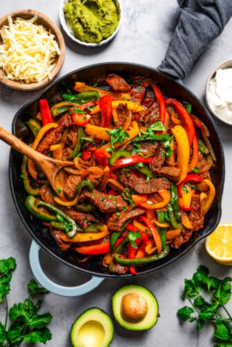Steak fajitas in a cast iron skillet with a wooden spoon surrounded by various ingredients.