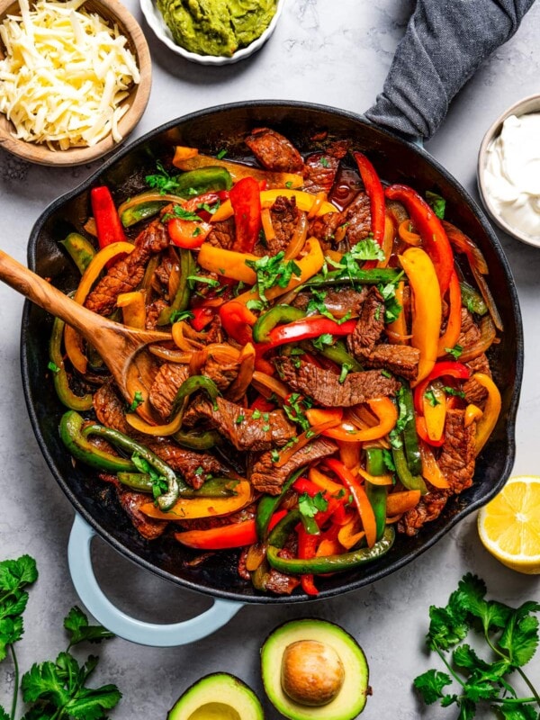 Steak fajitas in a cast iron skillet with a wooden spoon surrounded by various ingredients.
