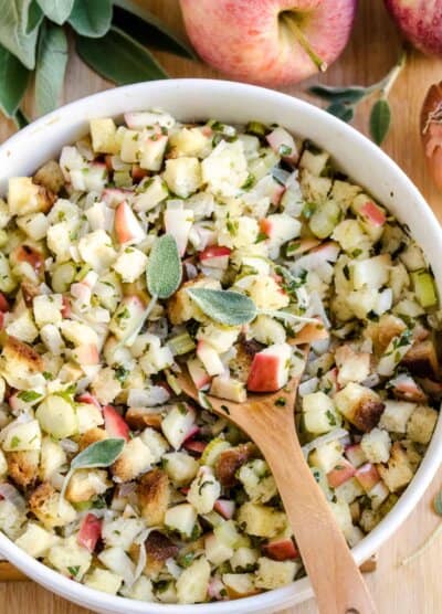 Apple stuffing in a white bowl with a wooden spoon topped with fresh sage