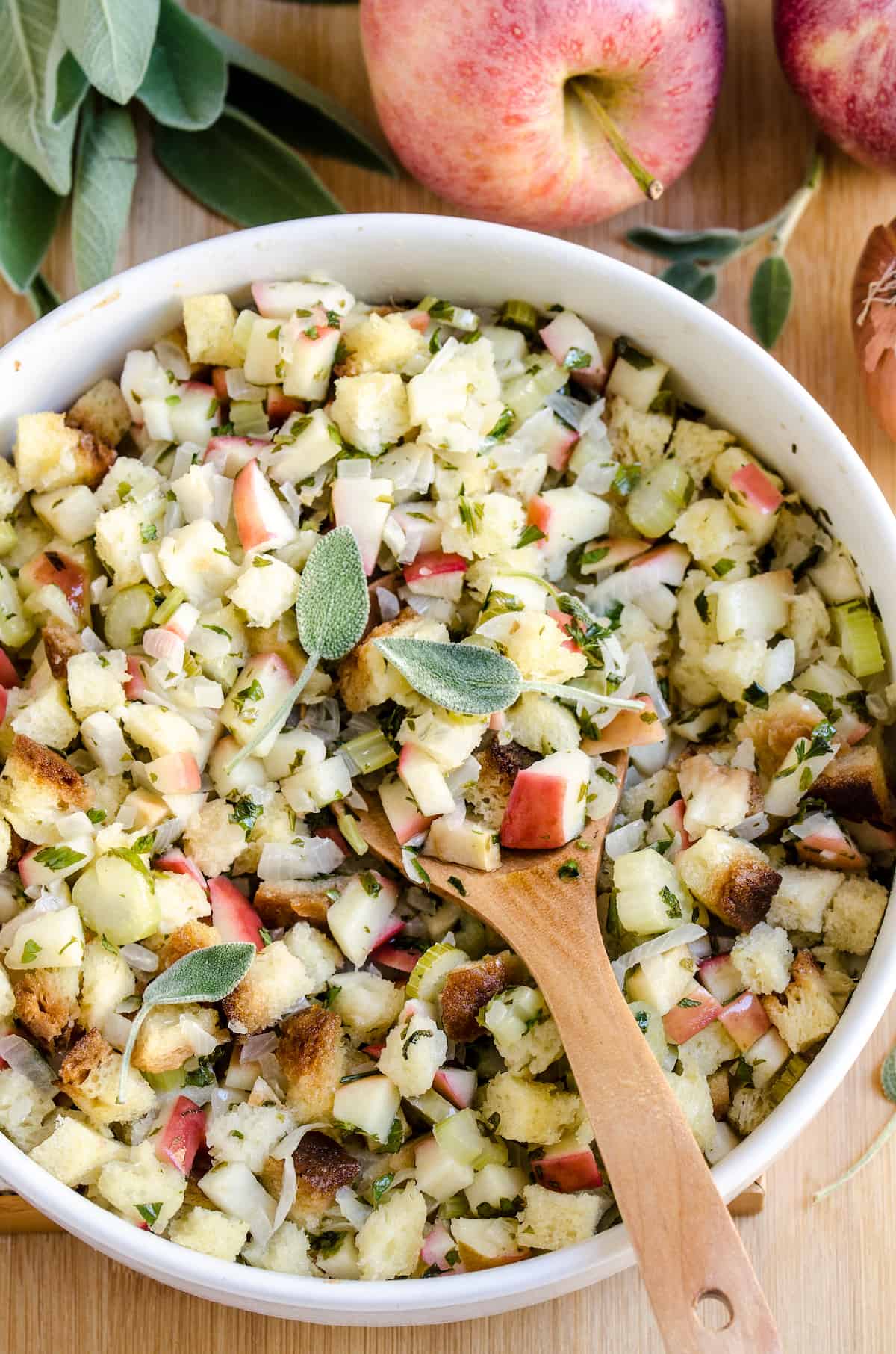 Apple stuffing in a white bowl with a wooden spoon topped with fresh sage
