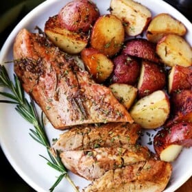 Partially sliced balsamic roasted pork tenderloin on a serving plate next to roasted potatoes and a sprig of rosemary