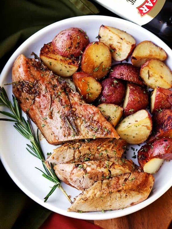 Partially sliced balsamic roasted pork tenderloin on a serving plate next to roasted potatoes and a sprig of rosemary
