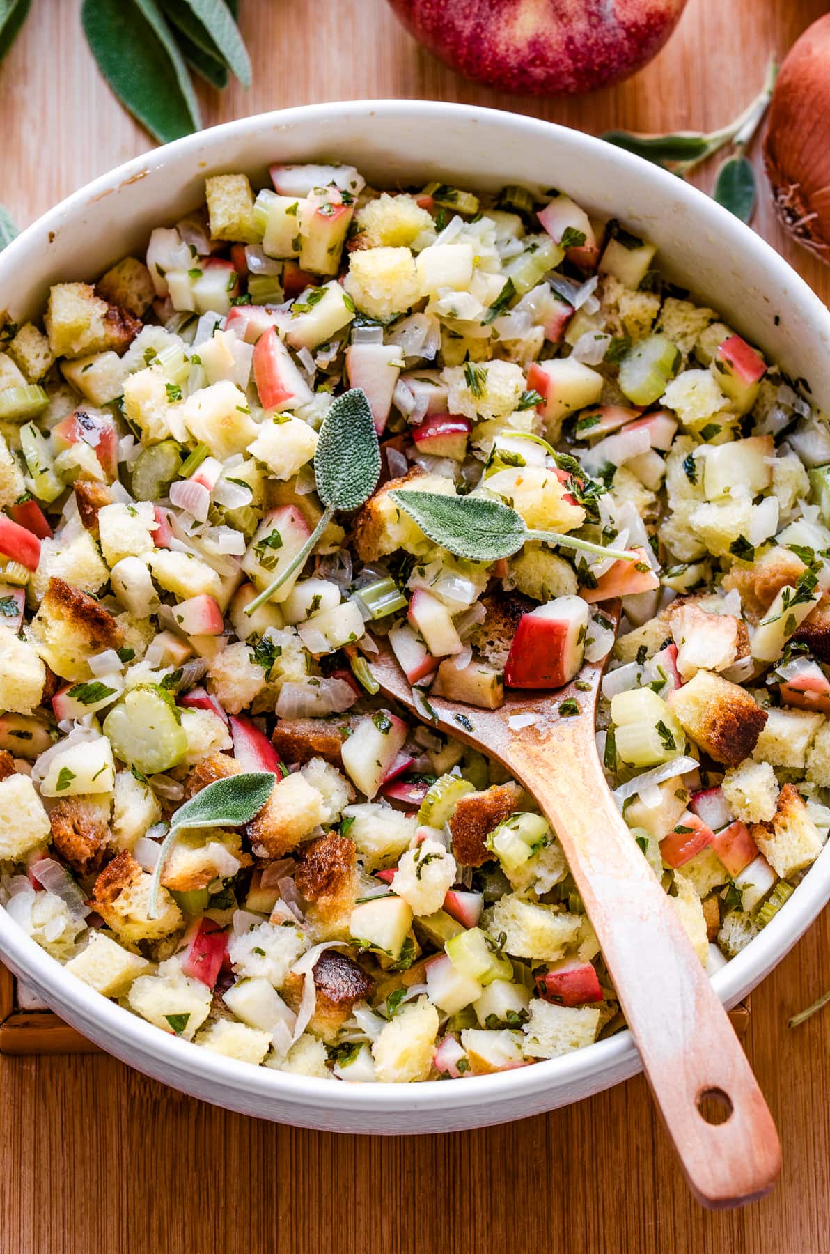 Apple stuffing in a white bowl with a wooden spoon inside the stuffing and topped with fresh sage.