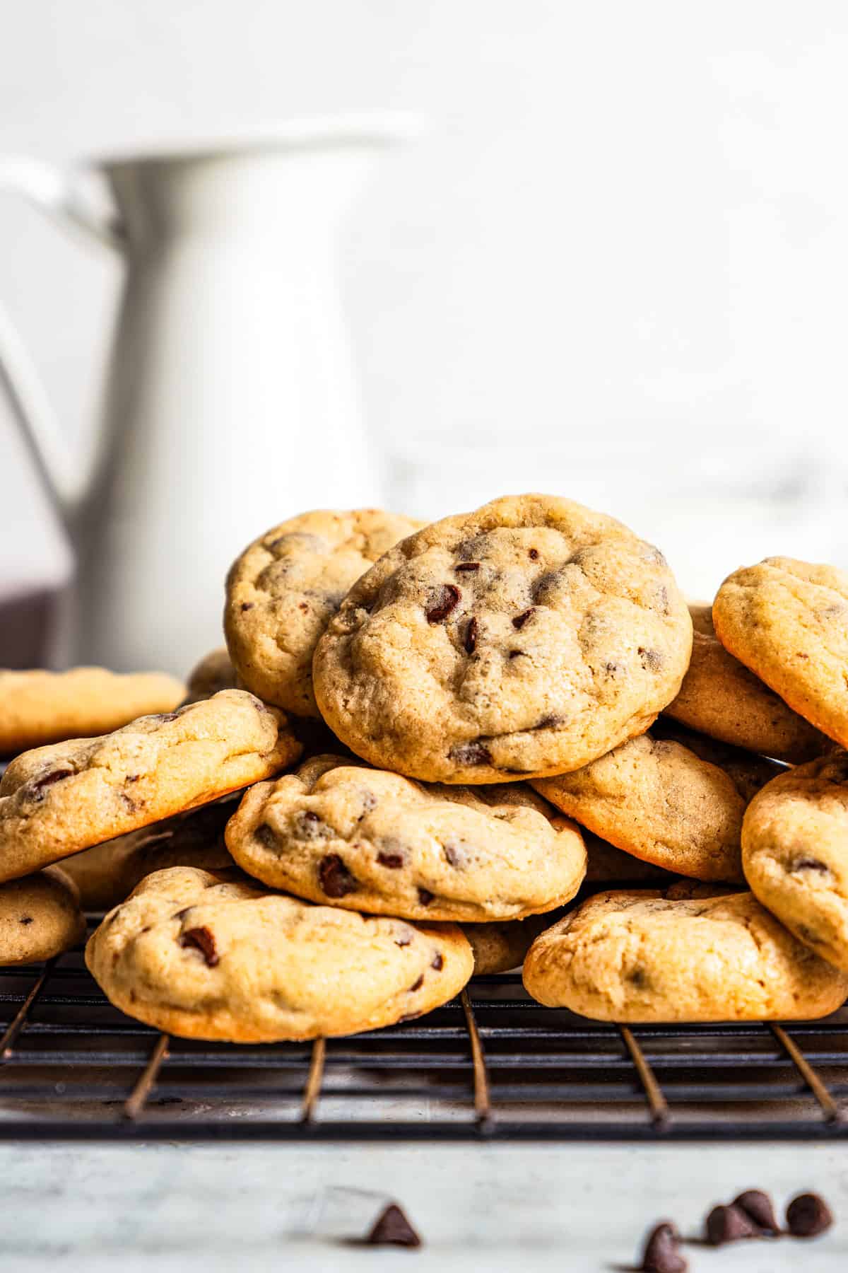 Mini chocolate chip cookies stacked on a wire rack with a ceramic milk jug in the background.