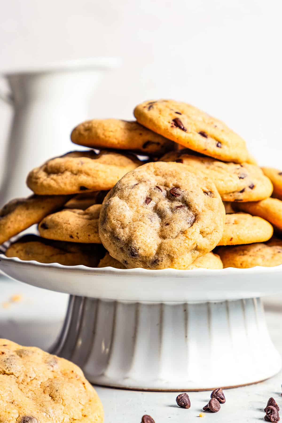 Mini chocolate chip cookies stacked on a wire rack with a ceramic milk jug in the background.